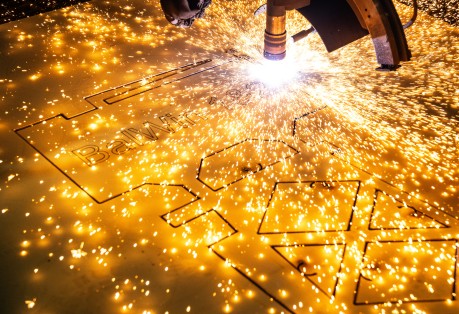 Das Bild zeigt eine Nahaufnahme eines Laserschneiders oder Plasmaschneiders, der in eine goldfarbene Metallplatte schneidet. Ein heller, funkelnder Lichtbogen und zahlreiche leuchtende Funken sprühen von der Schnittstelle. Das intensive Licht dominiert die rechte Seite des Bildes. Die Metallplatte ist horizontal ausgerichtet und weist bereits teilweise ausgeschnittene Formen auf, darunter der Text "BalWin". Der Hintergrund ist unscharf und dunkel, wodurch der Fokus vollständig auf den Schneidevorgang und die Funken gelegt wird. Die Szene vermittelt den Eindruck eines industriellen Fertigungsprozesses mit hoher Präzision und Energie.
English:
The image is a close-up shot of a laser or plasma cutter in action, cutting into a gold-colored metal sheet. A bright, sparkling arc and numerous glowing sparks emanate from the cutting interface. The intense light dominates the right side of the image. The metal plate is oriented horizontally and already shows partially cut-out shapes, including the text "BalWin". The background is blurred and dark, putting the entire focus on the cutting process and the sparks. The scene conveys the impression of an industrial manufacturing process characterized by high precision and energy.