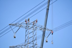 Drei Monteure in Schutzkleidung und Helmen arbeiten gesichert auf dem Querausleger eines Hochspannungsmastes in großer Höhe. Neben ihnen ist der Arm eines Krans zu sehen. Im Hintergrund ein wolkenloser blauer Himmel.