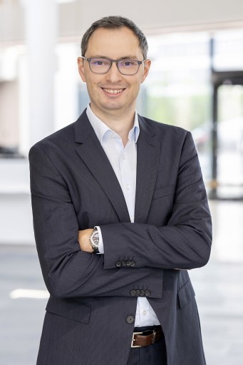 Ein Mann mittleren Alters mit Brille steht lächelnd in einem modernen Bürogebäude. Er trägt ein dunkles Sakko über einem hellen Hemd und hat die Arme locker verschränkt. Im Hintergrund ist der Eingangsbereich unscharf zu erkennen. Das Bild wirkt professionell und freundlich.