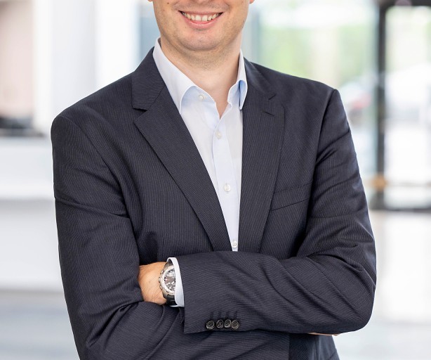 Ein Mann mittleren Alters mit Brille steht lächelnd in einem modernen Bürogebäude. Er trägt ein dunkles Sakko über einem hellen Hemd und hat die Arme locker verschränkt. Im Hintergrund ist der Eingangsbereich unscharf zu erkennen. Das Bild wirkt professionell und freundlich.