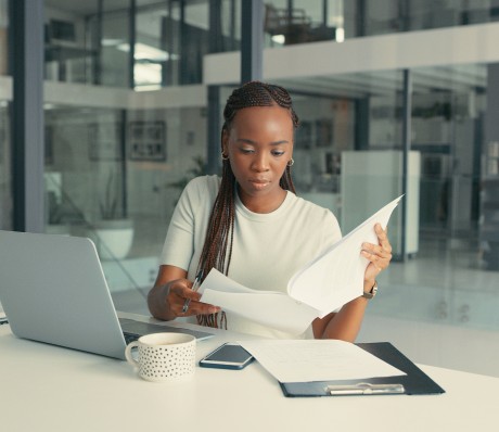 Eine Frau in Geschäftskleidung sitzt in einem gläsernen Büroraum. Vor ihr steht ein offener Laptop. Sie studiert konzentriert ein Dokument.
