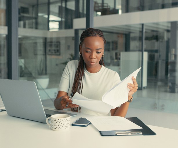 Eine Frau in Geschäftskleidung sitzt in einem gläsernen Büroraum. Vor ihr steht ein offener Laptop. Sie studiert konzentriert ein Dokument.