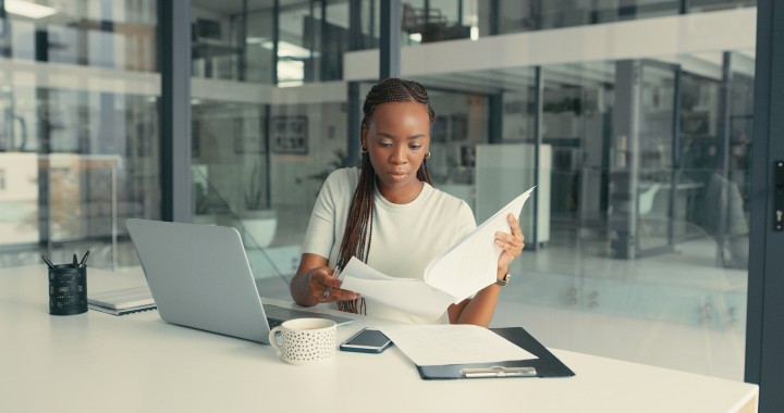 Eine Frau in Geschäftskleidung sitzt in einem gläsernen Büroraum. Vor ihr steht ein offener Laptop. Sie studiert konzentriert ein Dokument.