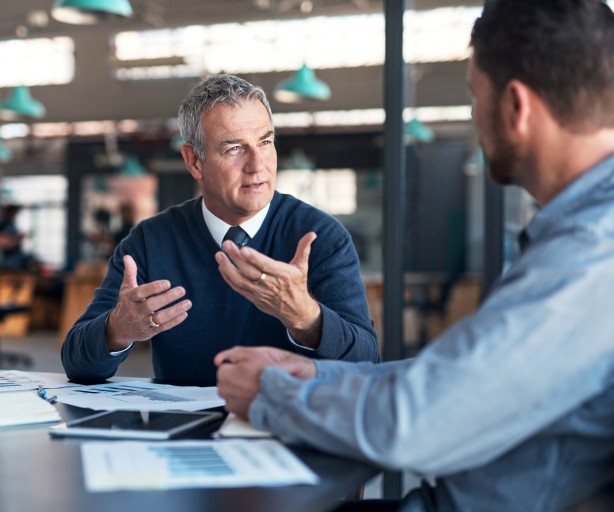 Zwei Männer in Geschäftskleidung sitzen einander zugewandt an einem Tisch in einem gläsernen Büroraum. Der Mann in der Bildmitte spricht und gestikuliert, auf dem Tisch liegen Unterlagen und ein Tablet.