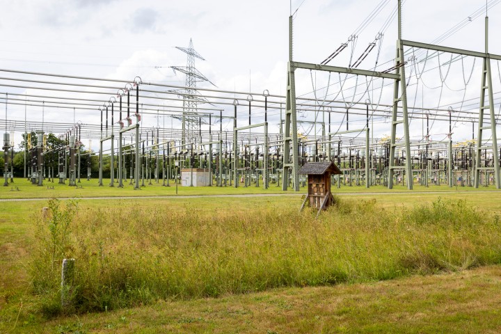 A sprawling substation facility under a cloudy sky. In the foreground, a green meadow with taller grass and some bushes extends. In the middle of the image, a small, wood-clad house with a pointed roof is visible. In the background, tall steel structures with numerous insulators and power lines running across the site dominate. A large overhead power line route with a transmission tower is visible on the horizon. The facility is surrounded by trees, which are visible on the left side of the image.