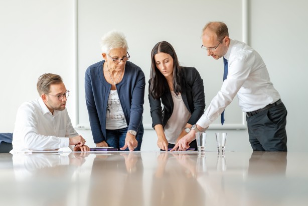 Das Foto zeigt zwei Männer und zwei Frauen. welche an einem Tisch stehen und auf Unterlagen blicken.
