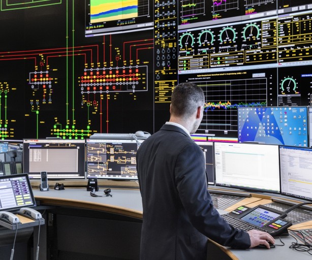 
Ein Kontrollraum mit großflächigen Monitoren, die Diagramme, Grafiken und Daten anzeigen. Zwei Männer in Anzügen sitzen oder stehen vor den Bildschirmen. Der Mann im Vordergrund ist von hinten zu sehen und bedient eine Tastatur. Der andere Mann sitzt weiter hinten links.