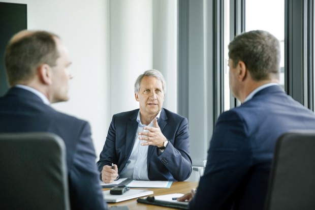 Drei Männer in Anzügen sitzen an einem Konferenztisch in einem modernen, hellen Besprechungsraum. Der Mann in der Mitte, mit grauen Haaren und einem dunkelblauen Sakko, spricht und gestikuliert leicht mit der rechten Hand. Er wirkt konzentriert und führt das Gespräch. Die beiden anderen Männer, deren Gesichter nur von der Seite zu sehen sind, hören aufmerksam zu. Der Raum hat große Fenster, die viel Tageslicht hereinlassen, und der Tisch ist mit Unterlagen, einem Tablet und Stiften bedeckt. Die Atmosphäre wirkt professionell und geschäftlich.
