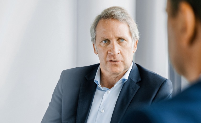 Ein Mann mit grauen Haaren und einem dunkelblauen Anzug sitzt in einem modernen, hellen Raum. Er schaut ernst und konzentriert in Richtung einer anderen Person, die unscharf im Vordergrund zu sehen ist. Die Person im Vordergrund trägt ebenfalls einen dunkelblauen Anzug. Der Hintergrund besteht aus großen, weißen Fenstern oder Wänden, was auf ein professionelles Gespräch in einem Büro hindeutet.