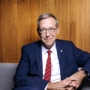 Ein Mann mittleren Alters mit kurzen grauen Haaren und Brille sitzt lächelnd vor einem holzvertäfeltem Hintergrund. Er trägt einen dunkelblauen Anzug, ein weißes Hemd und eine rote Krawatte mit feiner Struktur. Sein Ausdruck wirkt freundlich und professionell.