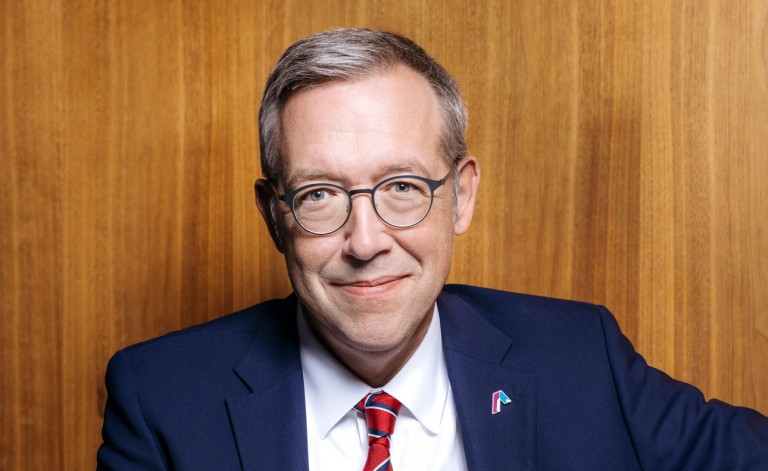Ein Mann mittleren Alters mit kurzen grauen Haaren und Brille sitzt lächelnd vor einem holzvertäfeltem Hintergrund. Er trägt einen dunkelblauen Anzug, ein weißes Hemd und eine rote Krawatte mit feiner Struktur. Sein Ausdruck wirkt freundlich und professionell.