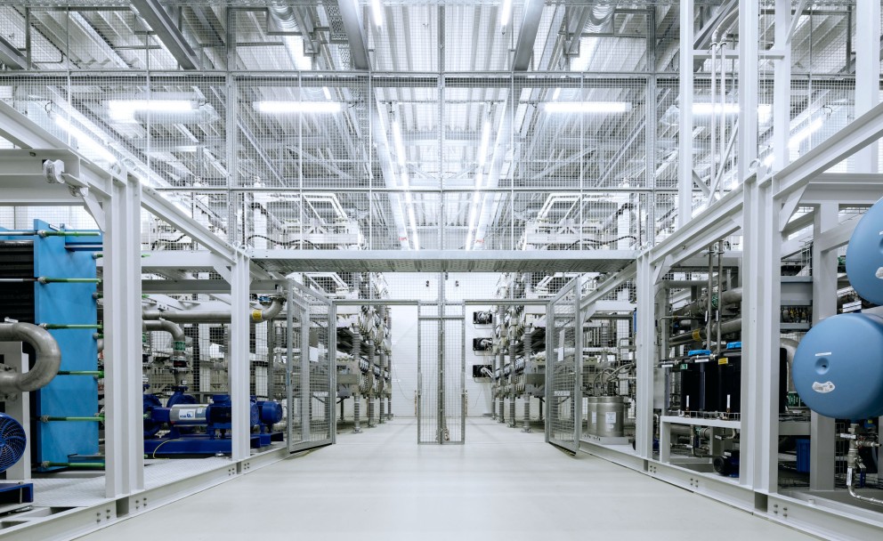 Das Bild zeigt eine STATCOM-Anlage (Static Synchronous Compensator) in einer industriellen Umgebung. Der Raum ist hell beleuchtet mit zahlreichen LED-Leuchten an der Decke. Die Anlage besteht aus symmetrisch angeordneten technischen Komponenten, wie Rohren, Ventilen, Kabeln und großen elektrischen Modulen. Rechts im Bild sind blaue, zylindrische Elemente zu erkennen, die Teil der Anlage sein könnten. Metallgitter trennen die Bereiche, und der Boden ist sauber und aus glattem, hellem Material. Die gesamte Konstruktion wirkt modern und hochfunktional, typisch für eine Einrichtung zur Regelung von Spannung oder Blindleistung im Stromnetz.