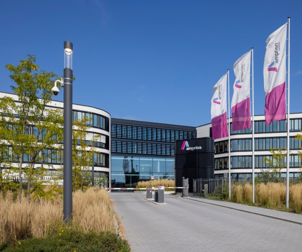Moderne Bürogebäudeanlage der Firma Amprion mit geschwungener Architektur und vielen Glasflächen. Im Vordergrund führt eine Einfahrt mit Schranke und Pollern auf das Gelände, das durch ein mittig gelegenes, dunkles Empfangsgebäude mit Amprion-Logo geprägt ist. Links und rechts davon erstrecken sich halbkreisförmige Gebäudeflügel mit weißen Fassaden und dunklen Fensterbändern. Drei Amprion-Fahnen in Weiß, Lila und Pink wehen am rechten Bildrand. Im Vordergrund sind Ziergräser, junge Bäume und eine Überwachungskamera an einem Mast sichtbar. Der Himmel ist klar blau.