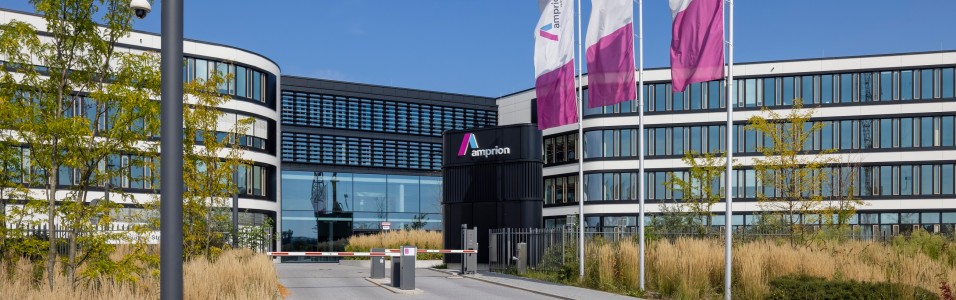 Moderne Bürogebäudeanlage der Firma Amprion mit geschwungener Architektur und vielen Glasflächen. Im Vordergrund führt eine Einfahrt mit Schranke und Pollern auf das Gelände, das durch ein mittig gelegenes, dunkles Empfangsgebäude mit Amprion-Logo geprägt ist. Links und rechts davon erstrecken sich halbkreisförmige Gebäudeflügel mit weißen Fassaden und dunklen Fensterbändern. Drei Amprion-Fahnen in Weiß, Lila und Pink wehen am rechten Bildrand. Im Vordergrund sind Ziergräser, junge Bäume und eine Überwachungskamera an einem Mast sichtbar. Der Himmel ist klar blau.