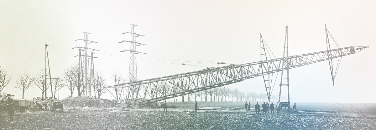 Historisches Bild vom Aufbau eines Strommasts. Der Mast wird schräg aufgestellt, umgeben von Arbeitern und landwirtschaftlicher Umgebung.