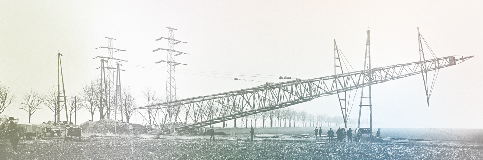 Historisches Bild vom Aufbau eines Strommasts. Der Mast wird schräg aufgestellt, umgeben von Arbeitern und landwirtschaftlicher Umgebung.