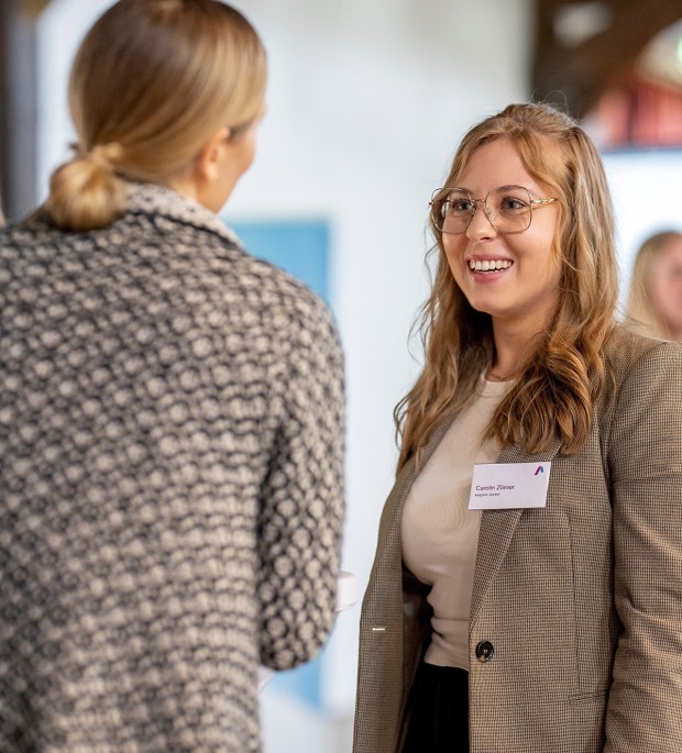 Auf dem Bild sind zwei Frauen zu sehen, die miteinander reden. Die rechte Frau ist von vorne zu sehen und lacht. Sie hat langes, braunes, leicht gewelltes Haar und trägt eine Brille mit einem hellen, filigranen Gestell. Sie hat einen Namensschild an ihrem braun-weiß gemusterten Blazer befestigt. Darunter trägt sie ein helles, einfarbiges Oberteil.
Die zweite Frau steht mit dem Rücken zur Kamera. Sie hat blonde Haare, die zu einem Pferdeschwanz gebunden sind, und trägt eine gemusterte Jacke in dunklen, unscharfen Farben. Der Hintergrund ist hell und unscharf, was den Fokus auf die Frauen lenkt.