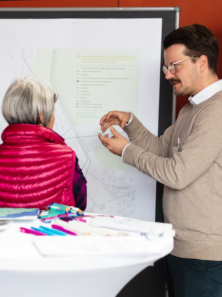 Vor einer Infotafel sind eine Besucherin und ein Mitarbeiter von Amprion im Gespräch. Der Mann macht eine erklärende Geste.