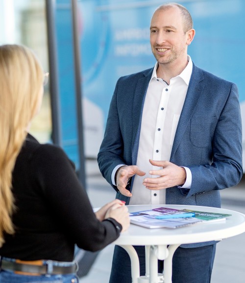 Auf dem Bild sind zwei Personen zu sehen, die an einem kleinen runden Tisch im Freien stehen und sich unterhalten. Der Fokus liegt auf einem Mann, der im Vordergrund steht und lächelt. Er ist hellhäutig, hat kurze Haare und trägt eine blaue Jacke über einem weißen Hemd mit dunklen Knöpfen. Er gestikuliert mit beiden Händen, während er spricht.
Von der zweiten Person, einer Frau, ist nur der Rücken zu sehen. Sie hat lange blonde Haare und trägt ein schwarzes Oberteil. Auf dem Tisch zwischen den beiden liegen einige Broschüren oder Hefte in verschiedenen Farben. Der Hintergrund ist unscharf und zeigt eine moderne Gebäudefassade in Blau- und Grautönen.