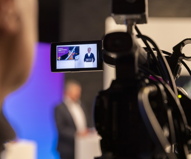 Das Foto zeigt eine Szene aus einem Film- oder Fernsehstudio. Im Vordergrund, links und unscharf, ist der Hinterkopf eines Menschen zu sehen, der wahrscheinlich die Kamera bedient. In der Mitte des Bildes dominiert eine professionelle Videokamera. Der aufgeklappte Monitor der Kamera zeigt eine Live-Aufnahme: Ein Mann im Anzug steht hinter einem Rednerpult und präsentiert etwas. Neben ihm auf dem Bildschirm ist eine Präsentationsfolie mit Grafiken zu sehen.
Im Hintergrund des Hauptbildes, ebenfalls unscharf, steht eine weitere Person in einem hell beleuchteten Bereich mit blau-lila Hintergrundbeleuchtung. Die Szenerie vermittelt den Eindruck einer professionellen Aufnahme für eine Pressekonferenz, einen Webcast oder eine ähnliche Veranstaltung.
