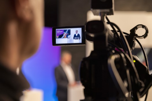 Das Foto zeigt eine Szene aus einem Film- oder Fernsehstudio. Im Vordergrund, links und unscharf, ist der Hinterkopf eines Menschen zu sehen, der wahrscheinlich die Kamera bedient. In der Mitte des Bildes dominiert eine professionelle Videokamera. Der aufgeklappte Monitor der Kamera zeigt eine Live-Aufnahme: Ein Mann im Anzug steht hinter einem Rednerpult und präsentiert etwas. Neben ihm auf dem Bildschirm ist eine Präsentationsfolie mit Grafiken zu sehen.
Im Hintergrund des Hauptbildes, ebenfalls unscharf, steht eine weitere Person in einem hell beleuchteten Bereich mit blau-lila Hintergrundbeleuchtung. Die Szenerie vermittelt den Eindruck einer professionellen Aufnahme für eine Pressekonferenz, einen Webcast oder eine ähnliche Veranstaltung.
