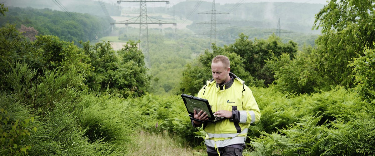 Ein Amprion-Mitarbeiter in gelber Schutzkleidung inmitten einer grünen Umwelt unter einer Freileitung