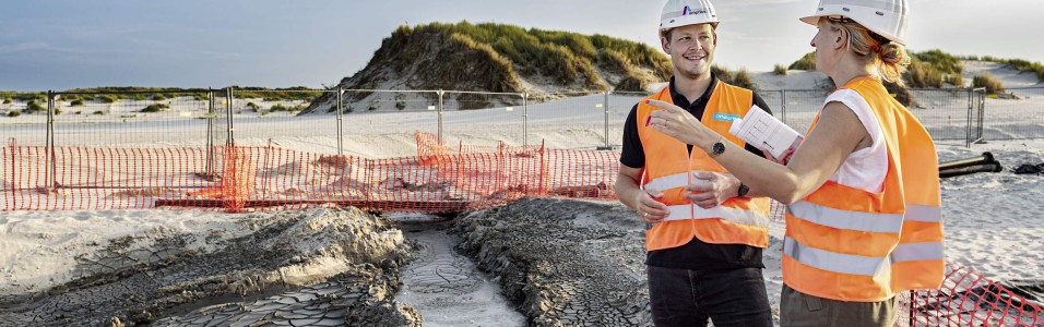 Zwei Amprion-Mitarbeitende in orangenen Warnwesten mit weißen Schutzhelmen auf dem Kopf stehen in einer Dünenlandschaft unter blauem Himmel. Alles ist mit Zaun und orangenen Netzen abgesperrt. Neben ihnen verschwindet ein rotes Rohr im Boden.