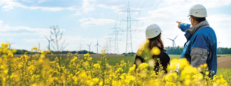 Das Titelbild des Nachhaltigkeitsberichts zeigt zwei Mitarbeiter vor einer Stromtrasse in einem Rapsfeld. Im Hintergrund sind Windräder zu sehen.