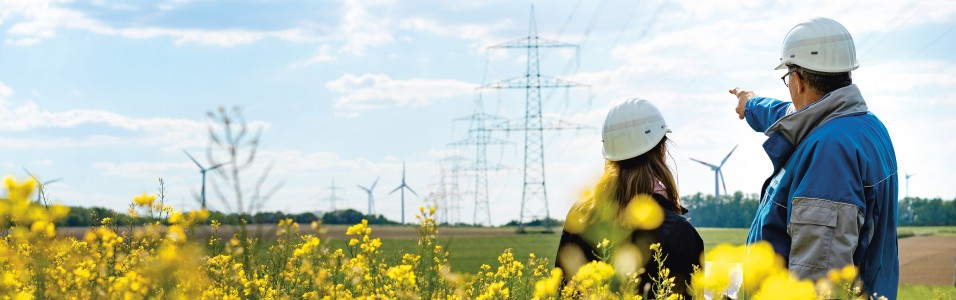 Das Titelbild des Nachhaltigkeitsberichts zeigt zwei Mitarbeiter vor einer Stromtrasse in einem Rapsfeld. Im Hintergrund sind Windräder zu sehen.