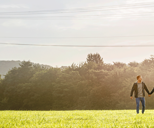Das Foto zeigt ein Pärchen Händchen haltenders Weise auf einer grünen Wiese, im Hintergrund ist Wald. Über Ihnen sind Höchstspannungsleitungen zu sehen. 