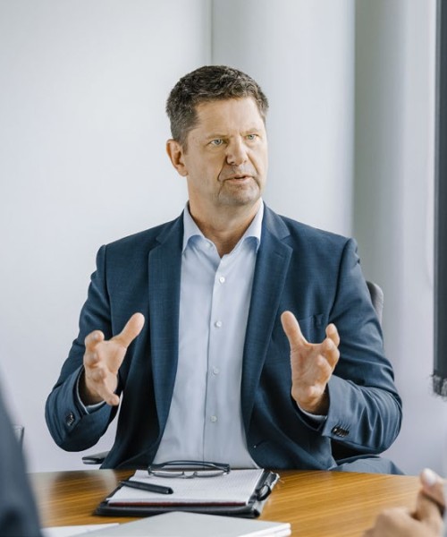 Das Bild zeigt den CFO der Amprion GmbH, Herrn Peter Rüth. Herr Rüth sitzt in einem Büro, vor ihm ein paar Unterlagen. Herr Rüth trägt ein dunkles Sakko über einem hellblauen Hemd ohne Krawatte. Er blickt den interviewenden an. Es ist eine klassische interviewsituation.