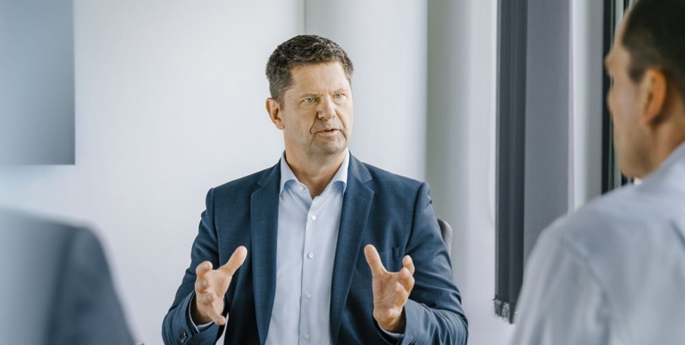 Das Bild zeigt den CFO der Amprion GmbH, Herrn Peter Rüth. Herr Rüth sitzt in einem Büro, vor ihm ein paar Unterlagen. Herr Rüth trägt ein dunkles Sakko über einem hellblauen Hemd ohne Krawatte. Er blickt den interviewenden an. Es ist eine klassische interviewsituation. 