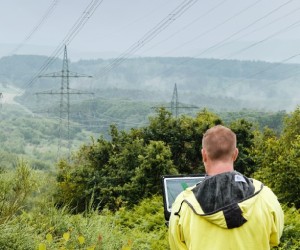 Ein Mann mit gelber Regenjacke steht mit dem Rücken zur Kamera in einer grünen, hügeligen Landschaft und betrachtet Hochspannungsmasten in der Ferne. Er hält ein Tablet oder einen Laptop in den Händen, vermutlich zur Inspektion oder Datenerfassung. Die Masten und Leitungen erstrecken sich durch die Landschaft und verschwinden im Nebel. Dichte Büsche und Bäume rahmen das Bild im Vordergrund ein, während das Wetter feucht und neblig erscheint.