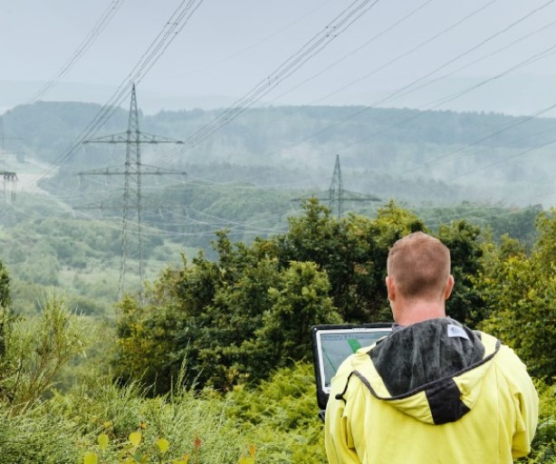 Ein Mann mit gelber Regenjacke steht mit dem Rücken zur Kamera in einer grünen, hügeligen Landschaft und betrachtet Hochspannungsmasten in der Ferne. Er hält ein Tablet oder einen Laptop in den Händen, vermutlich zur Inspektion oder Datenerfassung. Die Masten und Leitungen erstrecken sich durch die Landschaft und verschwinden im Nebel. Dichte Büsche und Bäume rahmen das Bild im Vordergrund ein, während das Wetter feucht und neblig erscheint.