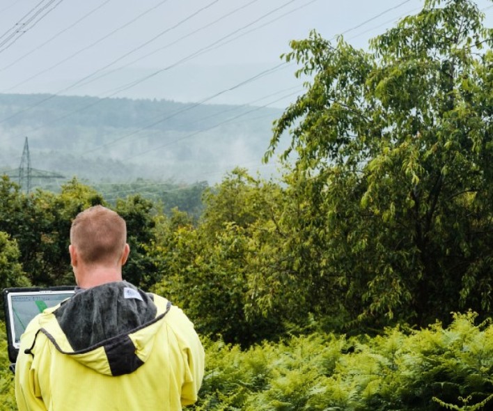 Ein Mann mit gelber Regenjacke steht mit dem Rücken zur Kamera in einer grünen, hügeligen Landschaft und betrachtet Hochspannungsmasten in der Ferne. Er hält ein Tablet oder einen Laptop in den Händen, vermutlich zur Inspektion oder Datenerfassung. Die Masten und Leitungen erstrecken sich durch die Landschaft und verschwinden im Nebel. Dichte Büsche und Bäume rahmen das Bild im Vordergrund ein, während das Wetter feucht und neblig erscheint.