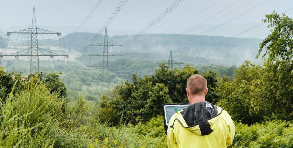 Ein Mann mit gelber Regenjacke steht mit dem Rücken zur Kamera in einer grünen, hügeligen Landschaft und betrachtet Hochspannungsmasten in der Ferne. Er hält ein Tablet oder einen Laptop in den Händen, vermutlich zur Inspektion oder Datenerfassung. Die Masten und Leitungen erstrecken sich durch die Landschaft und verschwinden im Nebel. Dichte Büsche und Bäume rahmen das Bild im Vordergrund ein, während das Wetter feucht und neblig erscheint.