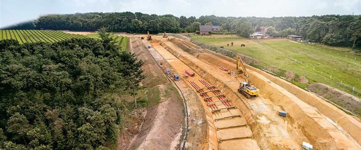 Luftaufnahme einer Baustelle mit Erdarbeiten und Baumaschinen neben einem Wald und landwirtschaftlichen Flächen.