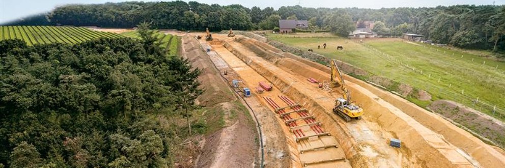 Luftaufnahme einer Baustelle mit Erdarbeiten und Baumaschinen neben einem Wald und landwirtschaftlichen Flächen.
