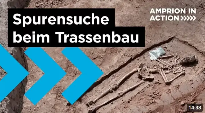 Das Bild ist ein Screenshot aus einem Video und zeigt eine Nahaufnahme eines vollständig freigelegten menschlichen Skeletts, das in einer archäologischen Grabungsfläche im lockeren Erdreich liegt. Der Fund wurde im Rahmen von Voruntersuchungen für den Bau der Amprion-Erdkabeltrasse A-Nord gemacht, wie der Text im Bild "Spurensuche beim Trassenbau" und "AMPRION IN ACTION" angibt. 
