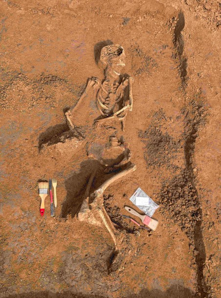 Menschliches Skelett in archäologischer Grabungsgrube, teilweise freigelegt, mit danebenliegendem archäologischem Werkzeug.