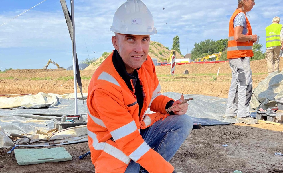 Archäologe in Sicherheitskleidung kniend an Ausgrabungsstätte.
