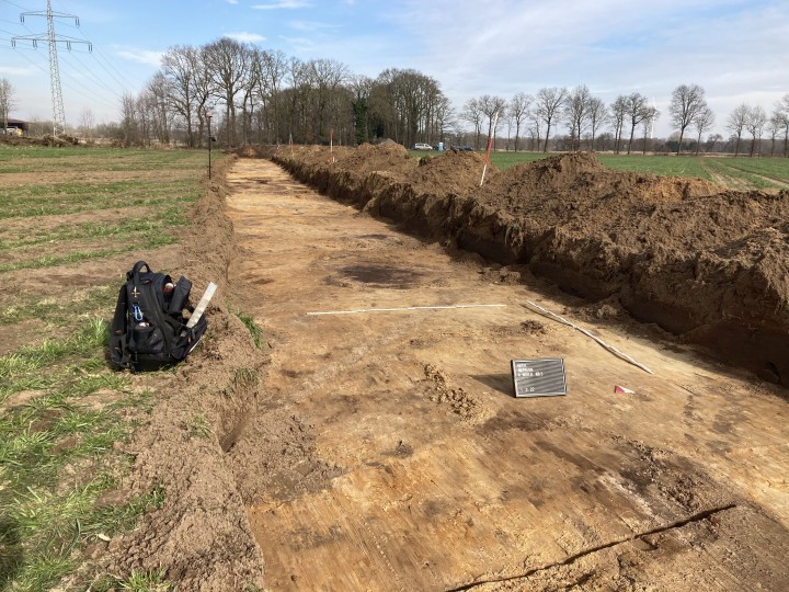 Baustelle: Länglicher archäologischer Grabungsschnitt in Agrarlandschaft, mit Werkzeug im Vordergrund.
