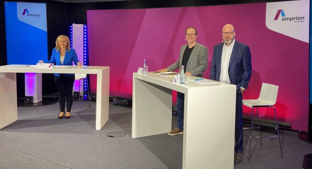 Drei Personen stehen lächelnd bei einer Amprion-Veranstaltung an Stehtischen auf der Bühne.
