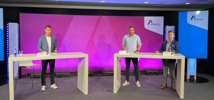 Drei Personen stehen lächelnd bei einer Amprion-Veranstaltung an Stehtischen auf der Bühne.
