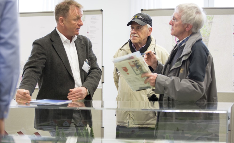 Drei Männer unterhalten sich an einem Tisch, im Hintergrund Präsentationstafeln.
