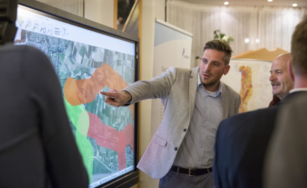 Mann im grauen Sakko zeigt mit dem Finger auf einen markierten Routenverlauf auf einem großen, beleuchteten Bildschirm. 