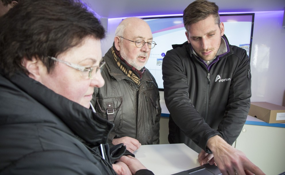 Drei Personen, die bei einer Veranstaltung stehen, betrachten gemeinsam einen Laptop. Eine Frau links und ein älterer Mann in der Mitte tragen Winterkleidung. Ein jüngerer Mann rechts in einer schwarzen Softshell-Jacke mit dem weißen Aufdruck "Amprion." zeigt mit dem Finger auf den Laptop-Bildschirm. Im unscharfen Hintergrund ist ein großer Bildschirm an der Wand erkennbar.