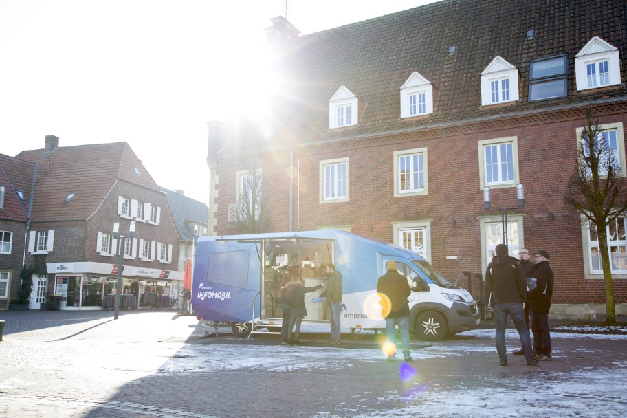 Ein blauer Kastenwagen, das sogenannte Infomobil des Stromnetzbetreibers Amprion, steht auf einem schneebedeckten Marktplatz, umgeben von historischen Backsteingebäuden.
Im Vordergrund des Bildes befinden sich mehrere Personen, die sich im Bereich der geöffneten Seiten des Fahrzeugs informieren oder unterhalten. Der Wagen trägt auf der Seite den Schriftzug "AMPRION" und "INFOMOBIL."