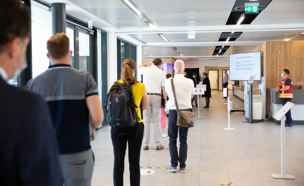 Warteschlange von Personen in einem modern beleuchteten Foyer, im Hintergrund Personal und Informationsbildschirme.