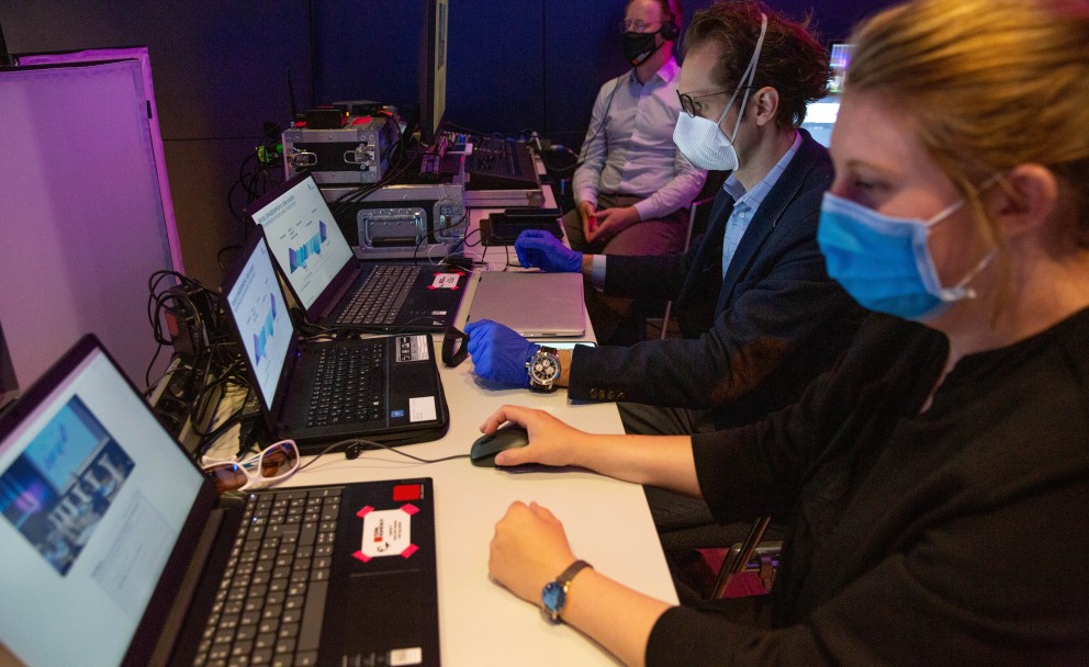 Drei Personen mit Masken arbeiten an Laptops in einem dunklen Raum.